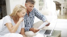 A couple look over paperwork together while sitting on the sofa in front of an open laptop.