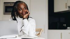 An older woman looks thoughtful as she sits at a kitchen table and looks into the distance.