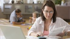 A woman works on her laptop and with a calculator while her husband and baby play in the background.