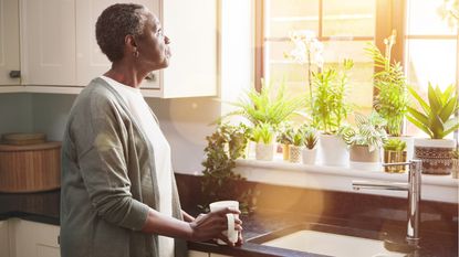 An older woman stands at the sink in her kitchen and thoughtfully looks out the window.