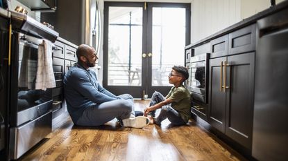 A father and and his young son sit on the floor in the kitchen and talk.