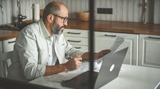 An older man looks at some paperwork while sitting in front of his laptop at his kitchen table.