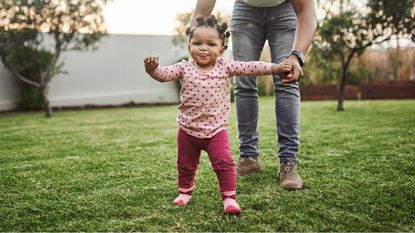 A toddler takes her first steps while outside on the lawn with her parent right behind her.