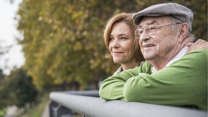 A young woman stands outside at a fence next to her aging father as they both look into the distance.