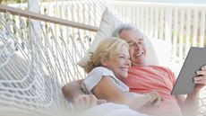 A happy older couple snuggle together in a hammock and look at a tablet together.