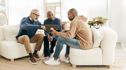 An older couple and their adult son look at a table together while sitting in the living room.